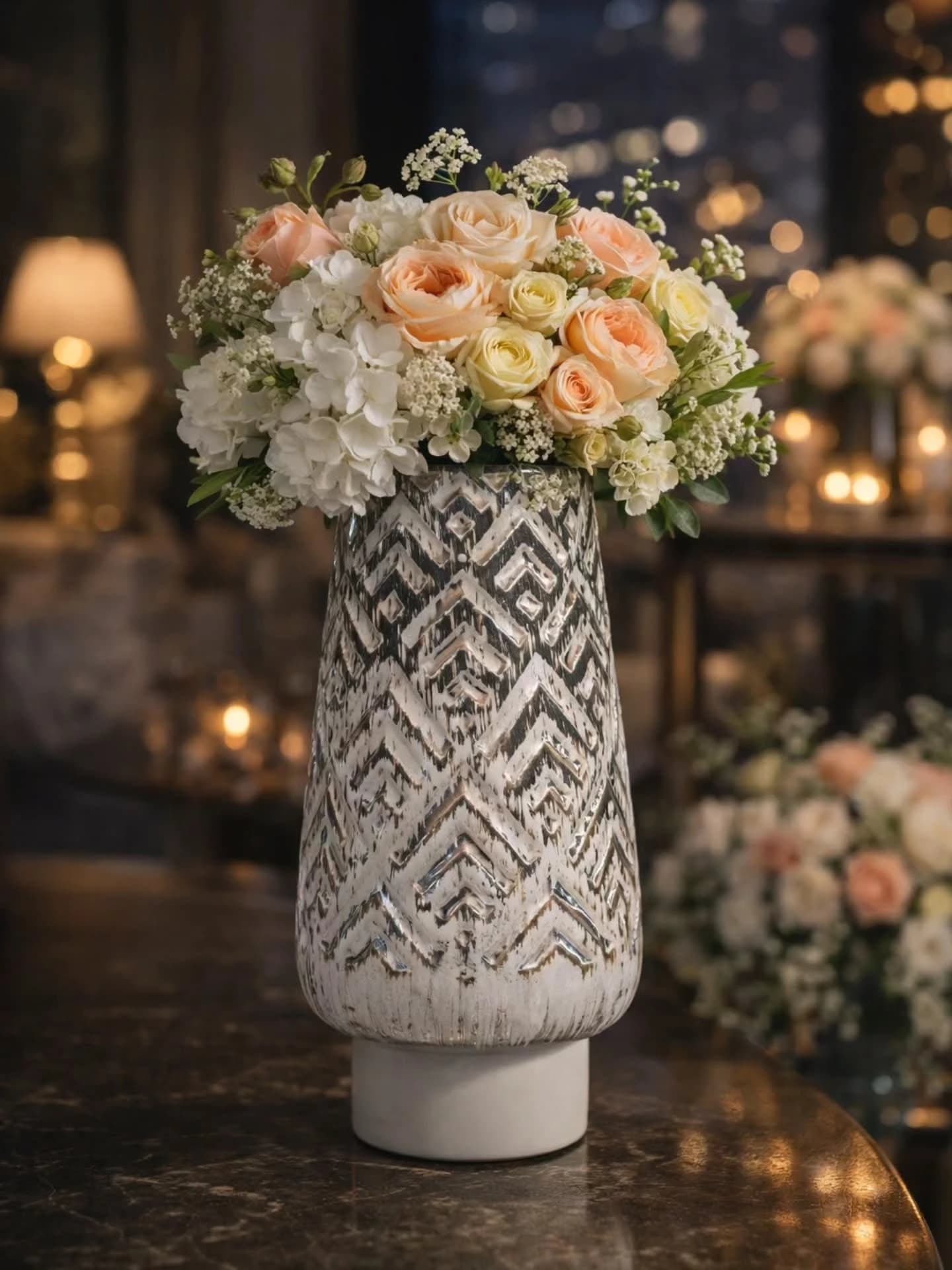 Tall ceramic vase with an embossed silver and white chevron pattern holding peach roses and white hydrangea, standing on a dark marble table with candles and window lights blurred behind.