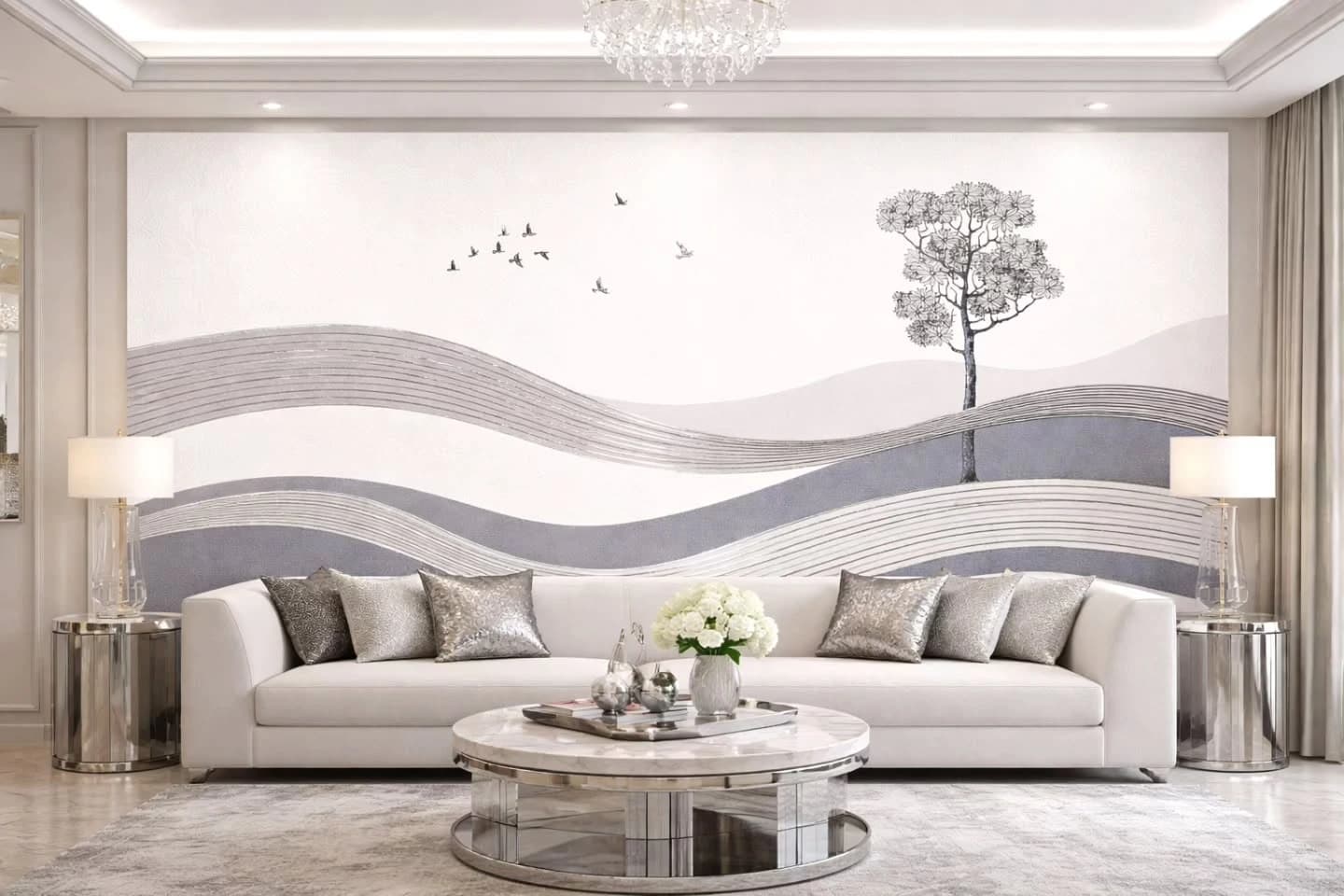 Formal living room with a long white sofa, silver textured pillows, round marble and chrome coffee table, matching lamps on mirrored side tables, and a full-width grey and white wave mural with a tree and flying birds behind it.
