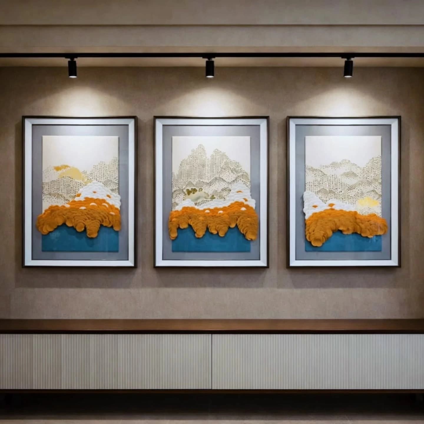 Three framed textured landscape artworks in orange, white and teal, lit by track spotlights above a white ribbed sideboard.
