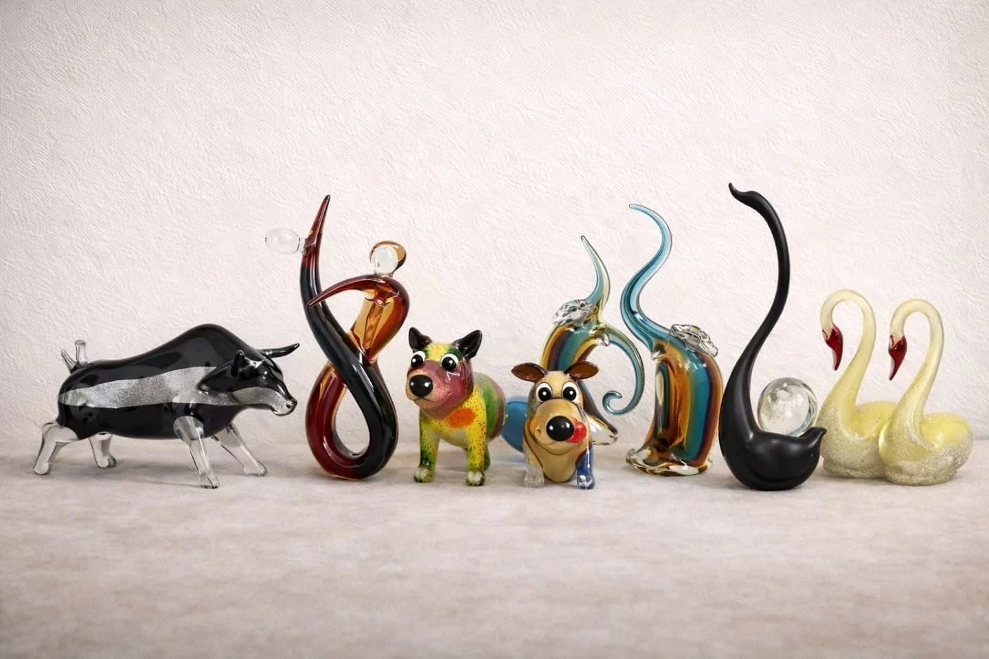 Eight blown-glass ornaments lined up against a plain off-white backdrop, including a black bull, a spotted dog, a cartoon cow, two abstract swirl forms, a black swan and a pair of pale swans.
