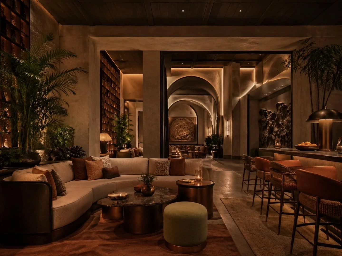 A warmly lit lounge with a curved cream sofa, bronze tables, planting and a bar counter with rattan stools beside a vaulted arched corridor.